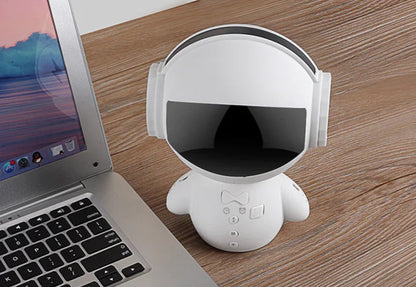 White robot-like device with headphones on a wooden desk next to a laptop.